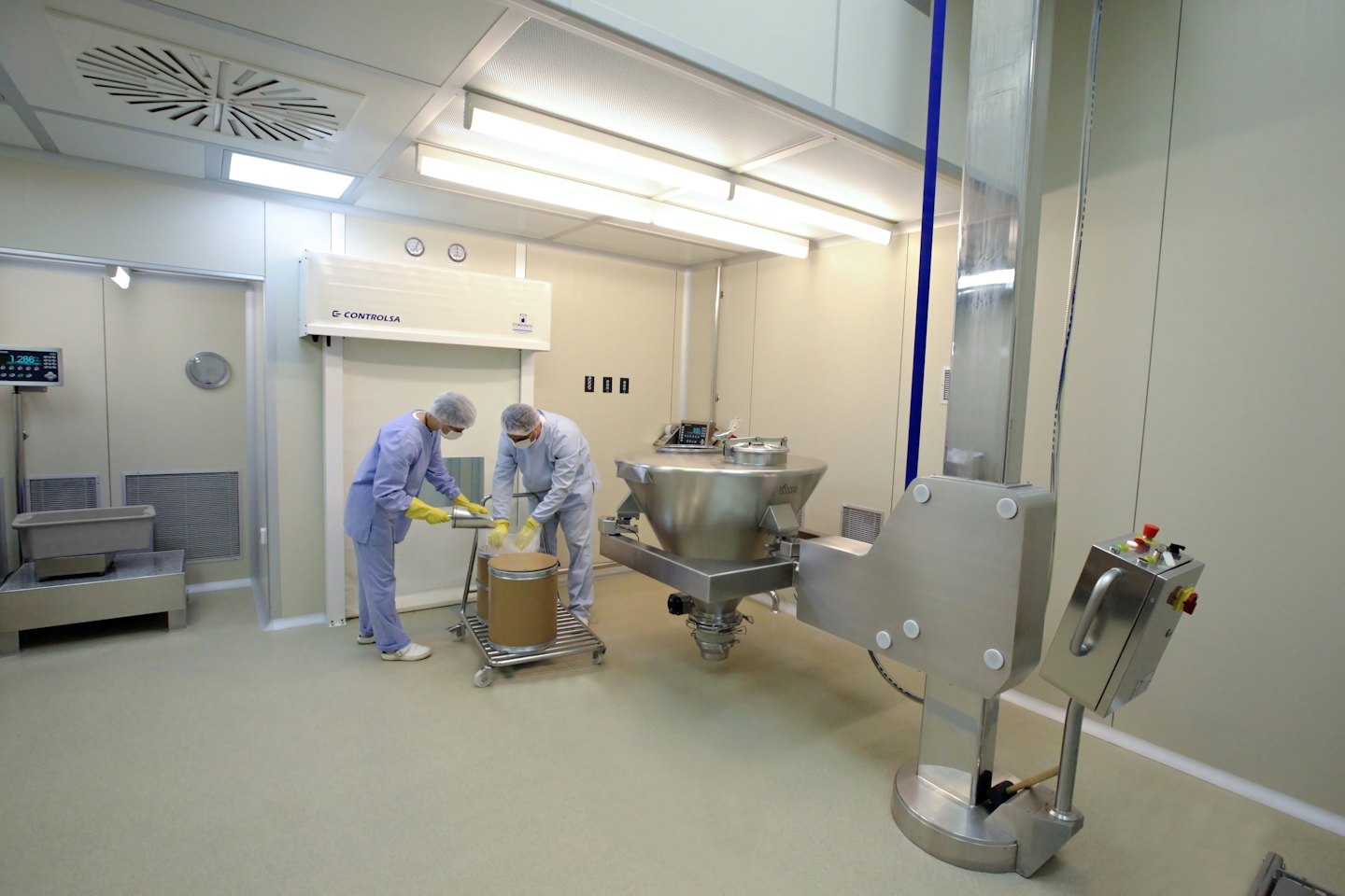 Two technicians in cleanroom suits pour material into a large drum.
