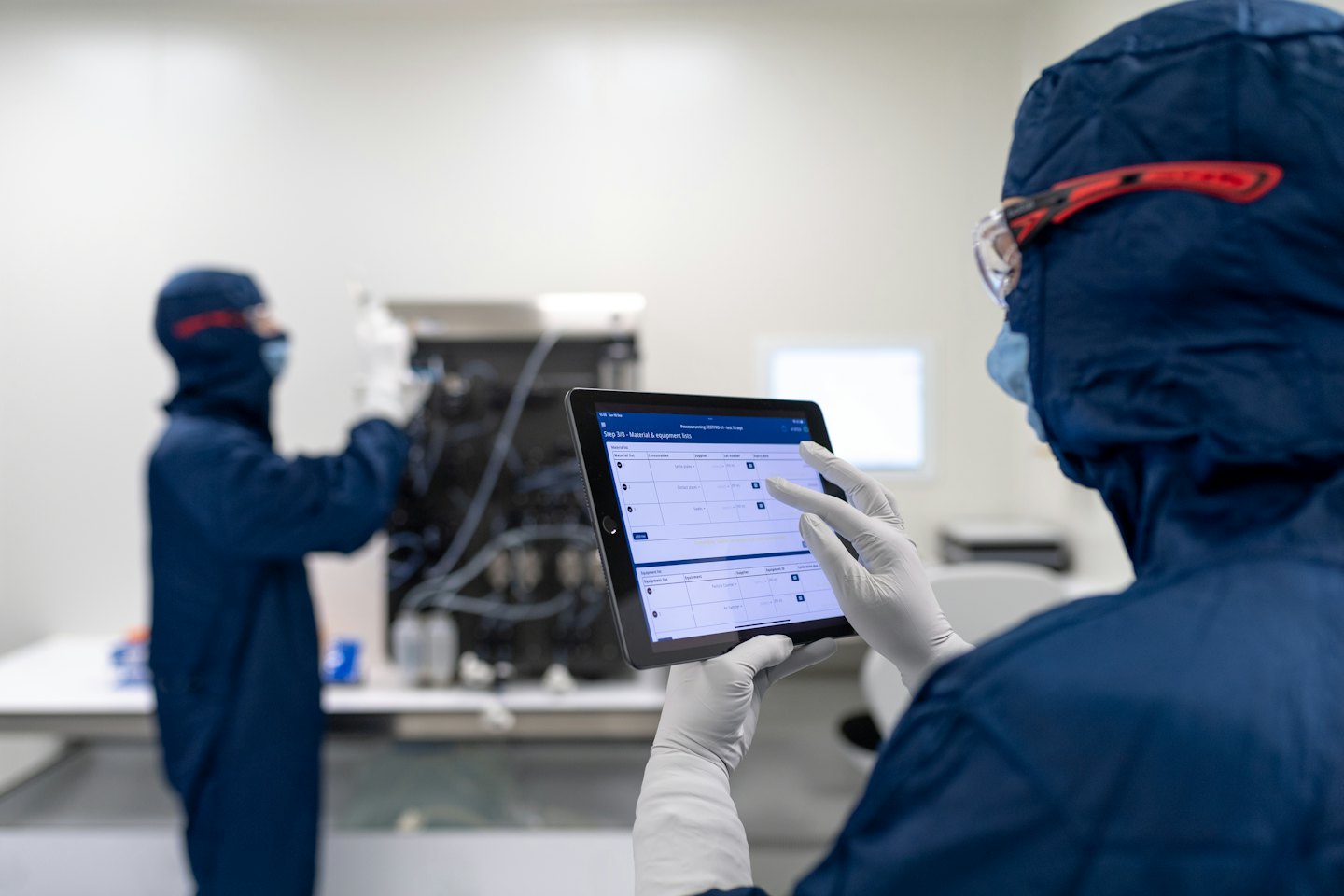 Scientists in cleanroom suits work with lab equipment and tablet.